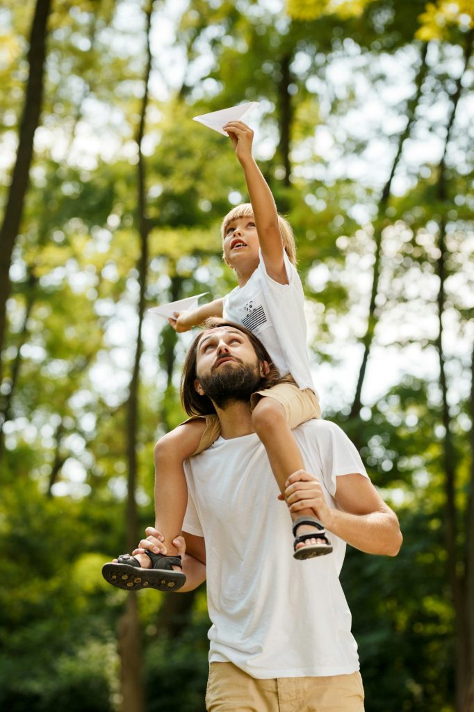 Walking in the forest. Blond boy dressed in a white t-shirt sitting on the shoulders of his bearded