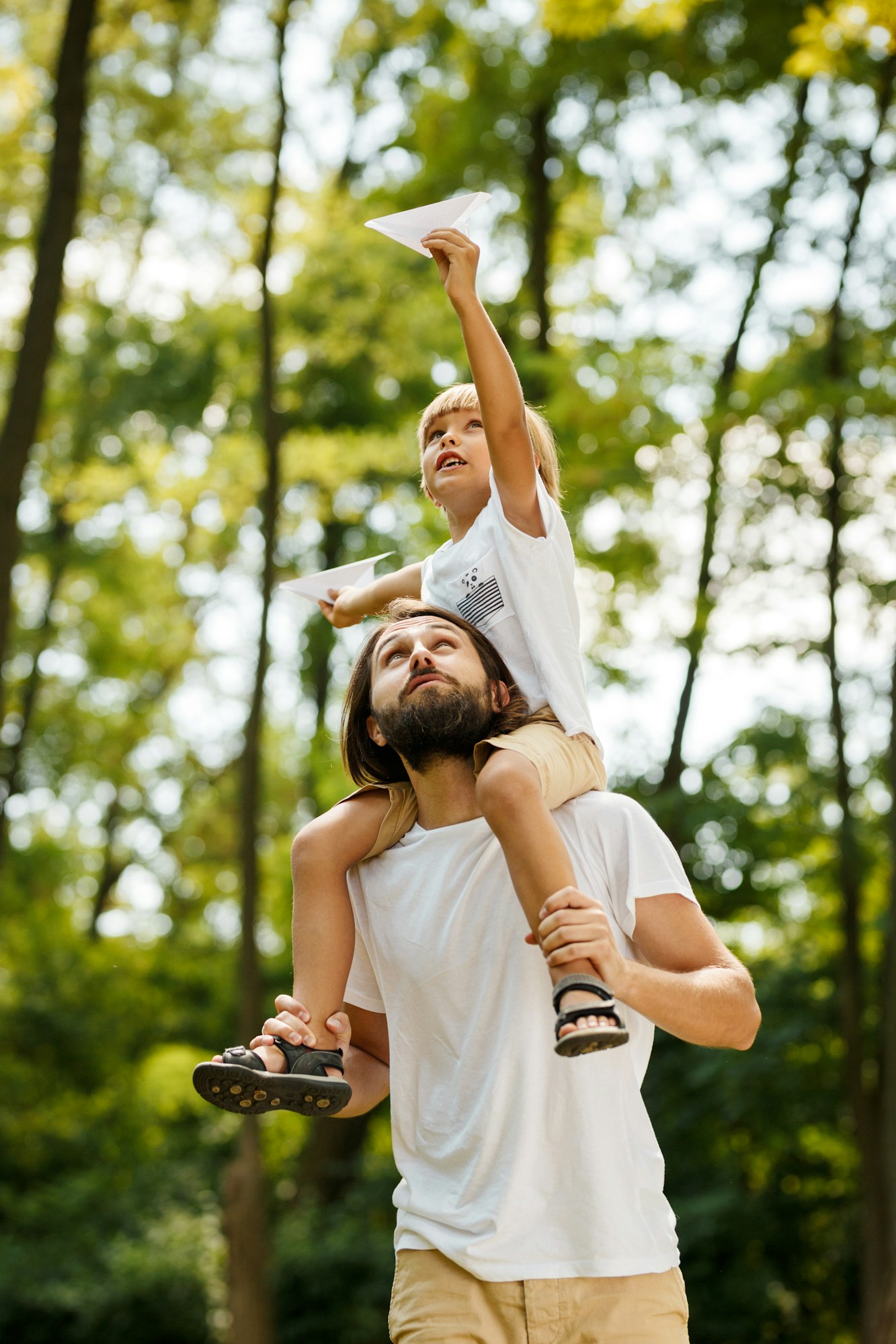 Walking in the forest. Blond boy dressed in a white t-shirt sitting on the shoulders of his bearded